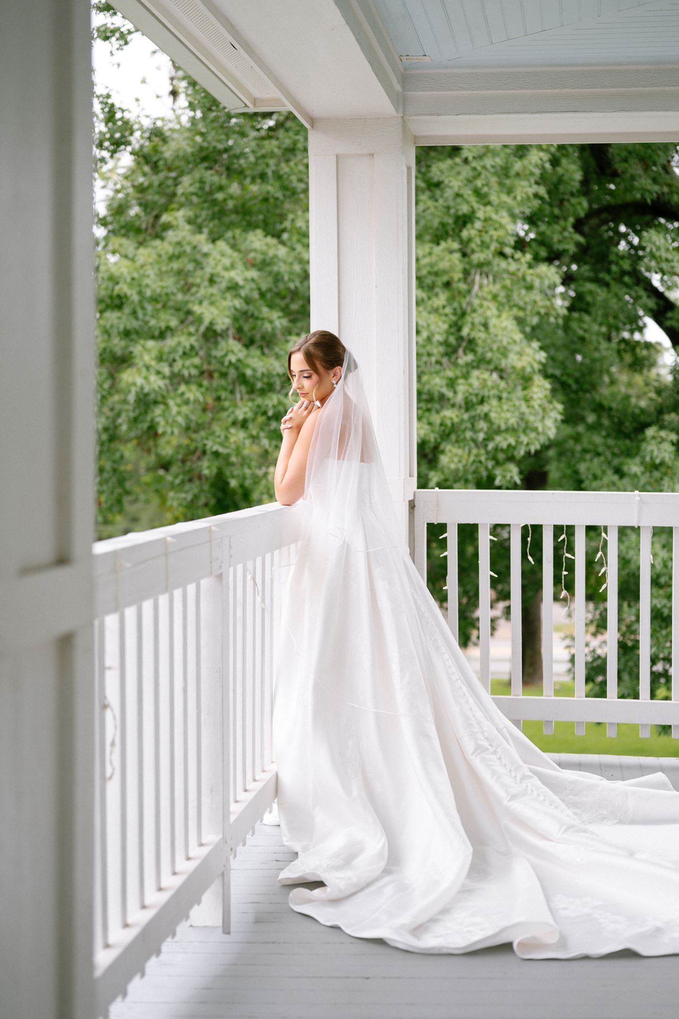 Bride in a flowing white gown and long veil stands on a white porch railing, surrounded by lush green trees.