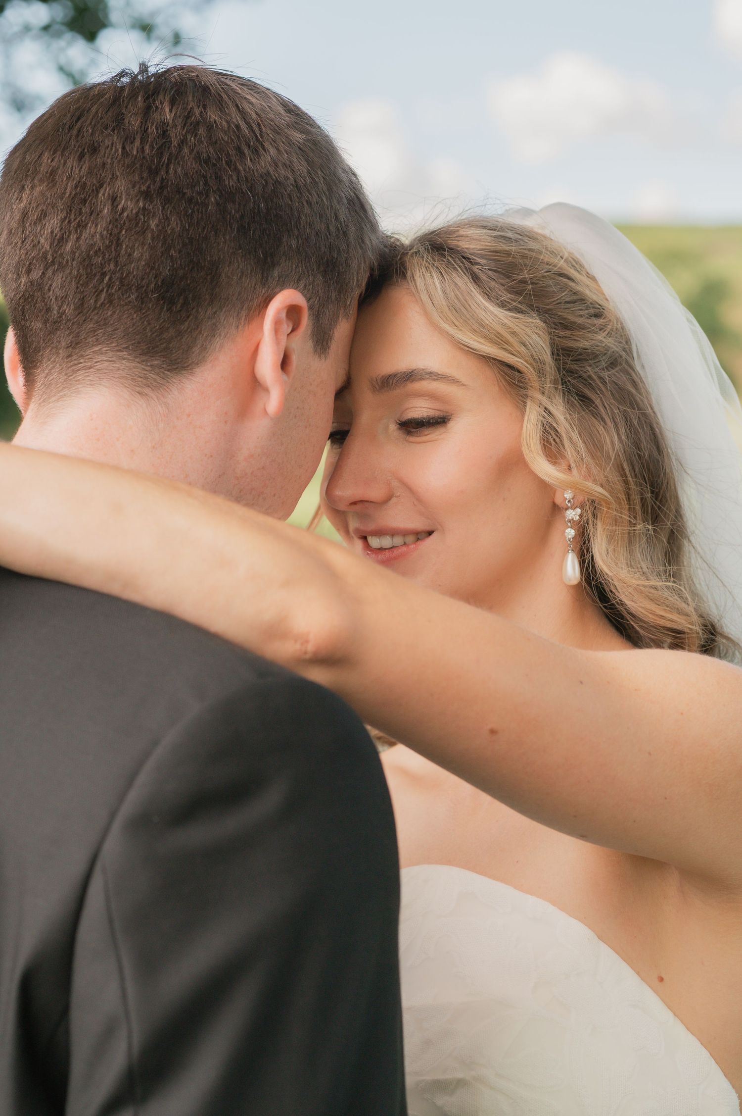 Bride and groom share an intimate forehead touch, bride smiling in white gown and veil with pearl earrings outdoors.