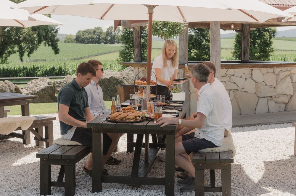 People dining al fresco at a rustic wooden table under white umbrellas at a scenic vineyard.