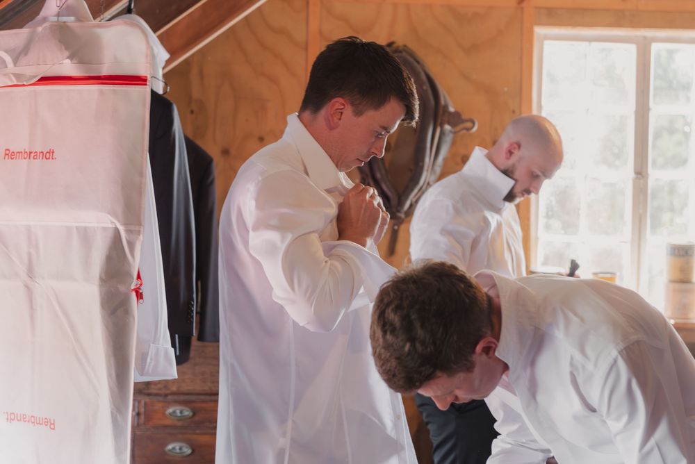 Men in white dress shirts getting ready, adjusting ties in a wooden room with hanging garments.