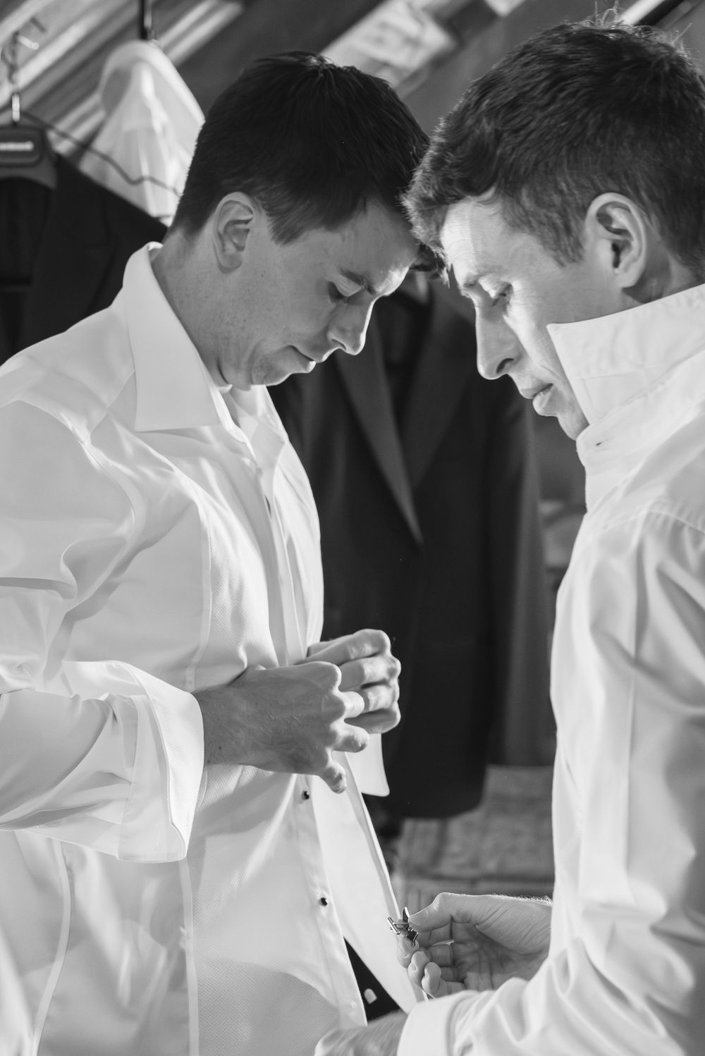 Two men in white dress shirts helping each other button up before a formal wedding ceremony.