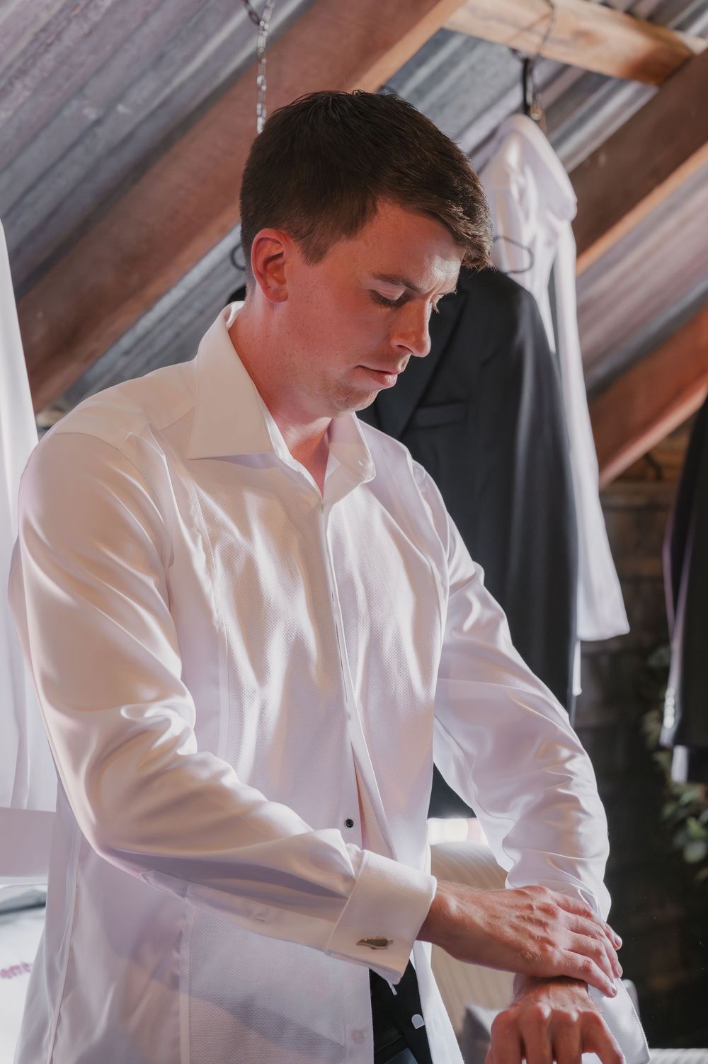 A man in a white dress shirt adjusts his cufflinks while getting dressed for a formal event.