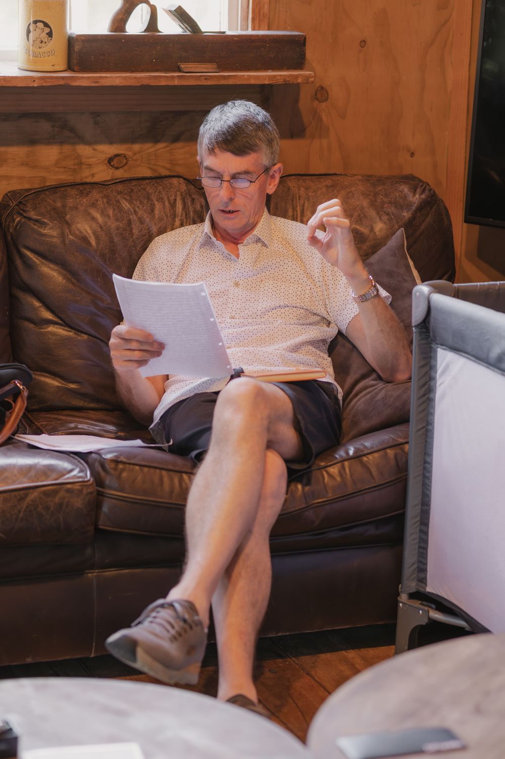 Older man with glasses reading documents while seated in a brown leather armchair indoors.
