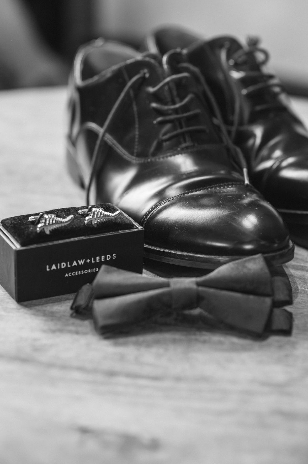 Black and white photo of polished oxford dress shoes, bow tie, and Laidlaw Leeds cufflinks box on a marble surface.