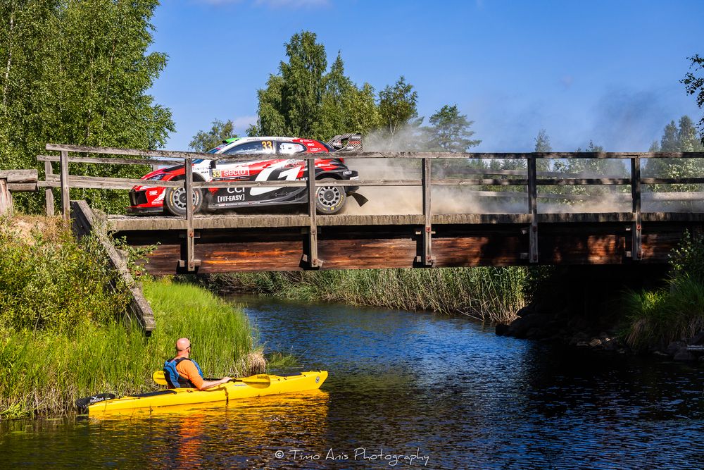 Rally car launches off a wooden bridge over a calm river while a kayaker watches from below on a sunny day.