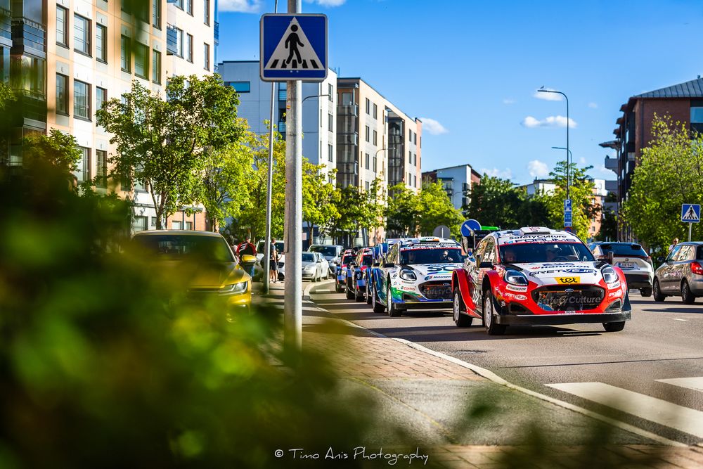 Rally cars race through a sunny urban street lined with trees and a pedestrian crossing sign during a WRC event.