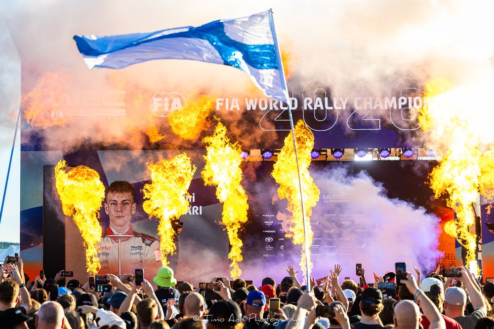 FIA World Rally Championship celebration with flames, Finnish flag, and cheering crowd at awards ceremony.