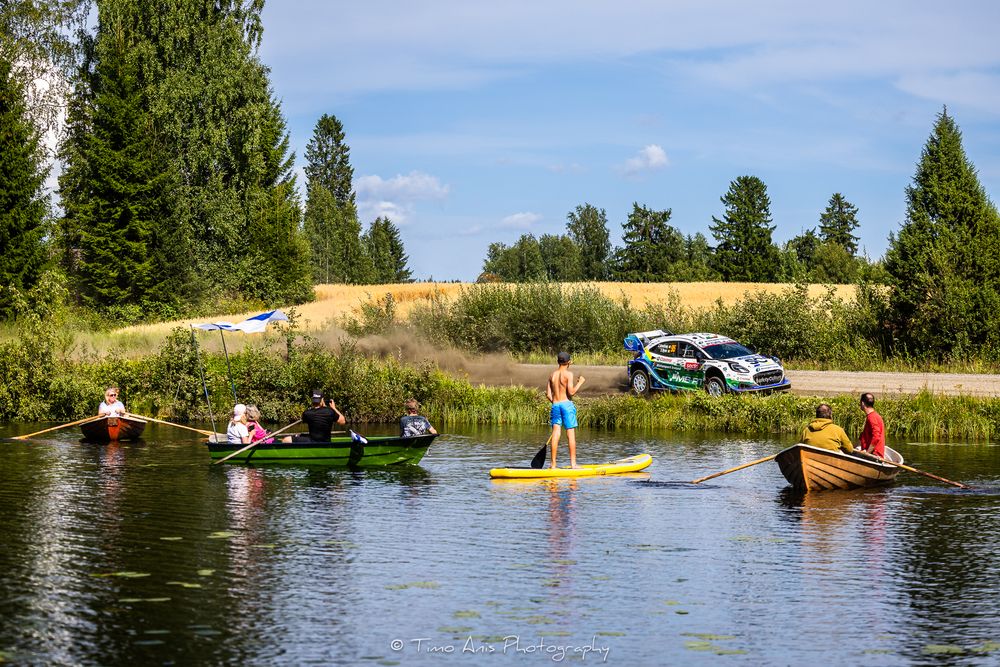 People kayaking and paddleboarding on a calm river with a rally car racing past on a nearby road in a scenic landscape.