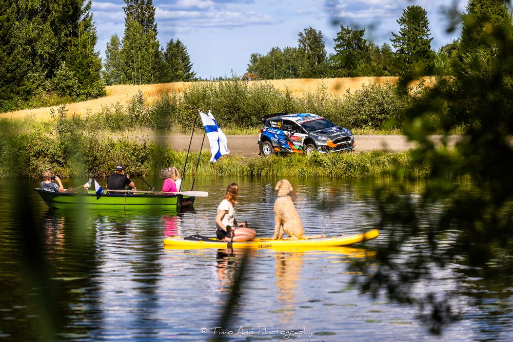 A rally car races past spectators kayaking and canoeing on a calm Finnish lake surrounded by lush summer greenery.