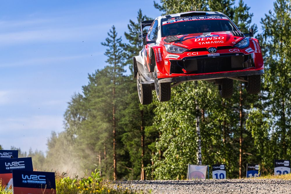 A red Toyota WRC rally car soars through the air during a jump on a forest stage rally course.