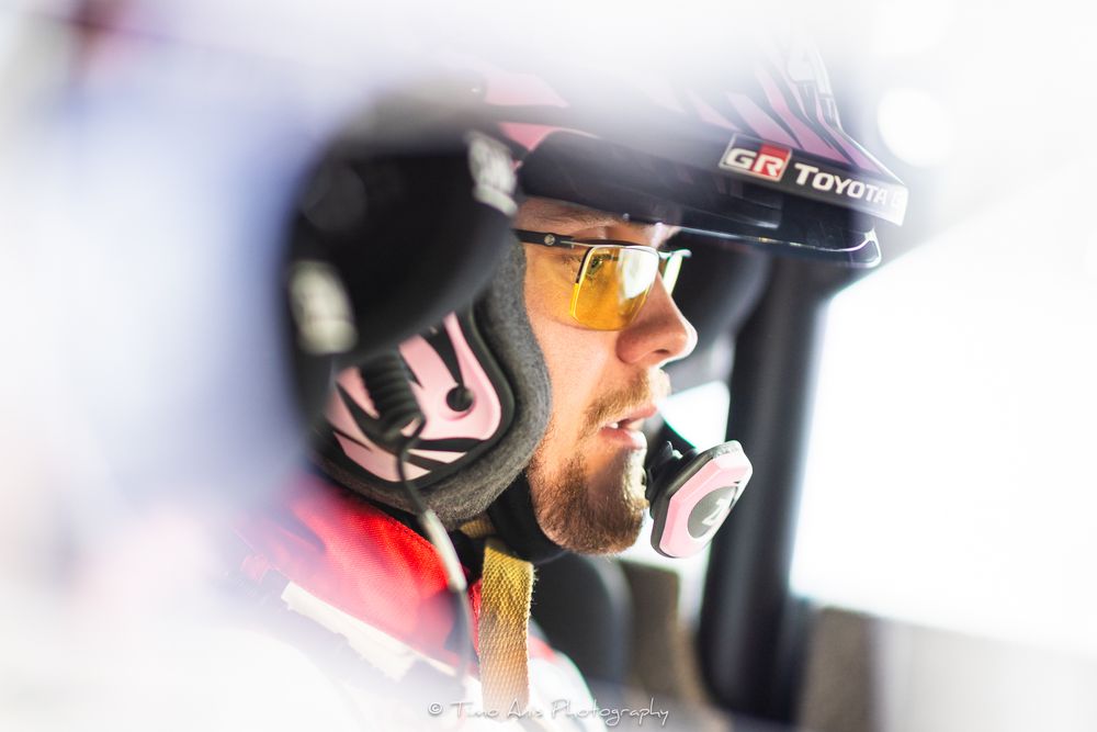 A focused male rally driver wearing a GR Toyota cap and pink earbuds sits in a race car cockpit.