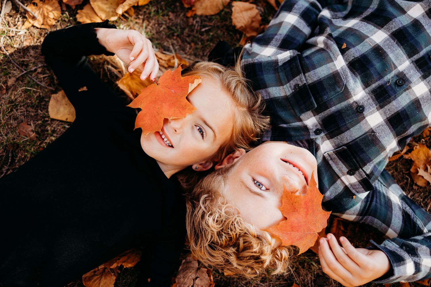 A brother and sister laying down beside each other on the ground with a leaf over one of their eyes smiling. There are leaves on the ground.