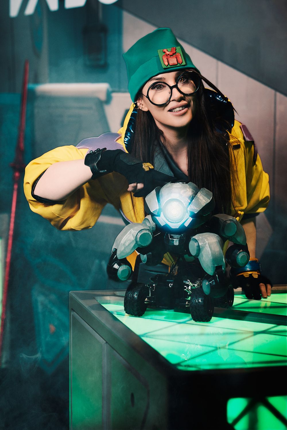Woman in yellow jacket and green hat cosplaying with a glowing robot prop in a neon-lit sci-fi setting.
