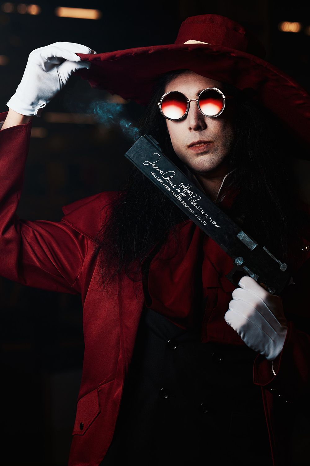 Person cosplaying in red coat and hat with round red glasses, holding a prop gun, resembling Alucard from Hellsing.