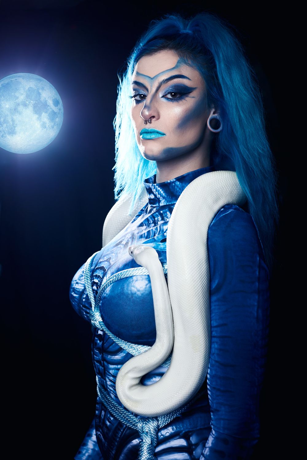 Woman in blue snake-themed costume with dramatic makeup holds a white python under a full moon.