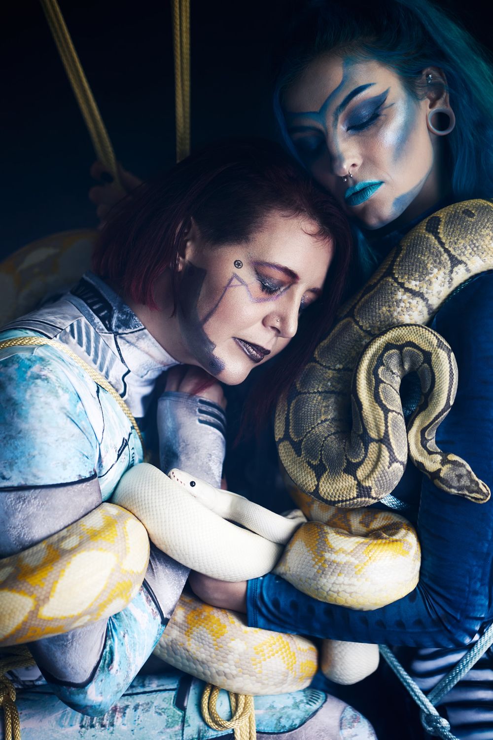 Two women with elaborate body paint pose with large snakes in a dramatic, artistic dark fantasy photo shoot.