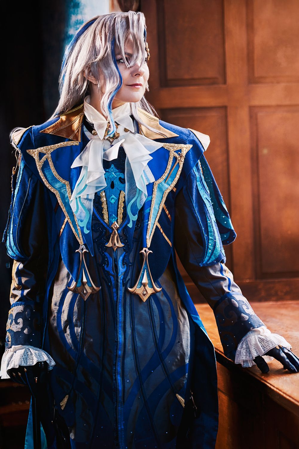 A cosplayer in an elaborate blue and gold fantasy costume with a white ruffled collar and ornate details.
