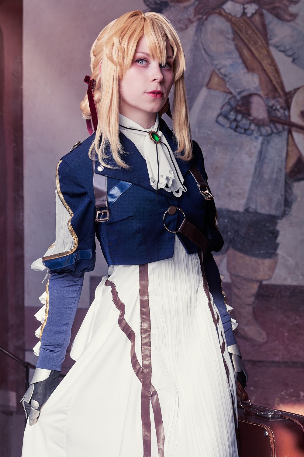 Cosplayer in Violet Evergarden costume with blonde wig, blue military jacket, and white dress holding a suitcase.