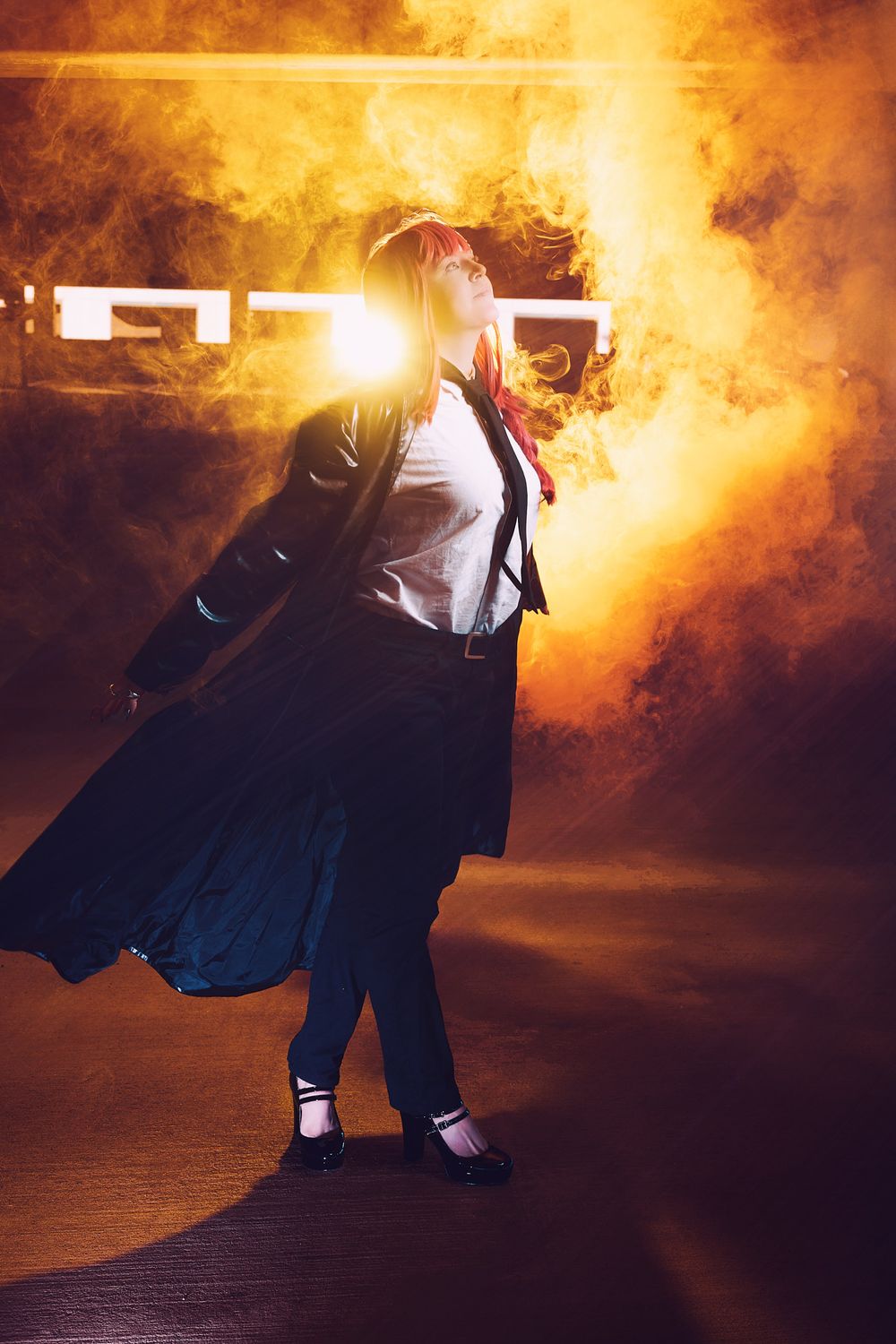 A cosplayer in a dark trench coat poses dramatically against a fiery orange smoke backdrop with glowing light effects.