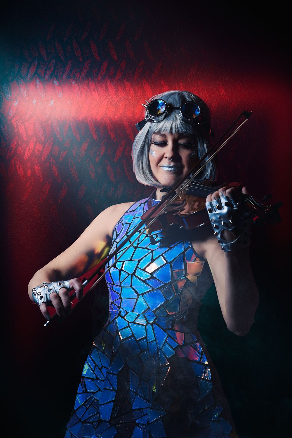 Woman in mosaic dress and steampunk goggles playing violin on dramatic red and blue lit stage.