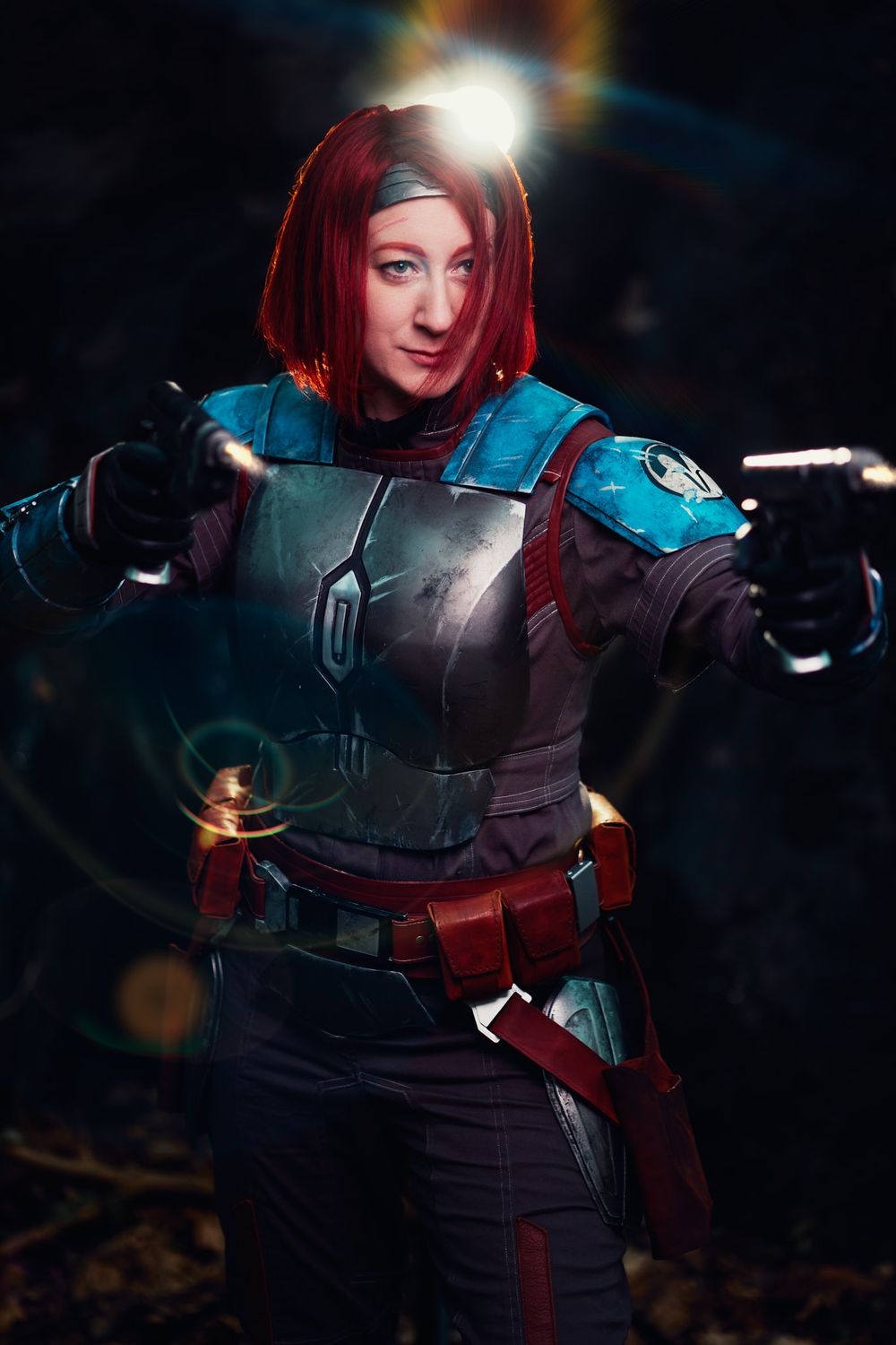 Woman in blue and red Mandalorian armor cosplay holding two blasters in a dramatic dark forest setting.