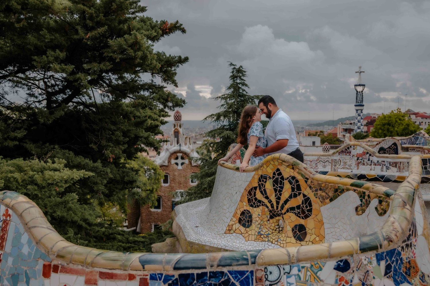 A couple posing for their engagement photoshoot sitting on the famous colorful mosaic benches of Park Güell, overlooking the Barcelona city