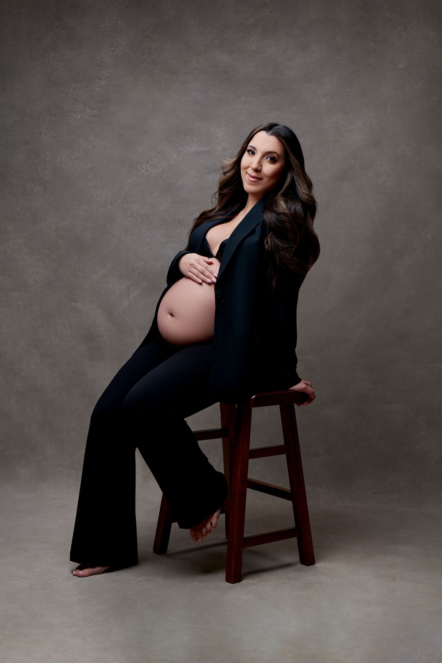Pregnant woman in black outfit sitting on wooden stool, cradling baby bump, studio maternity portrait photo.