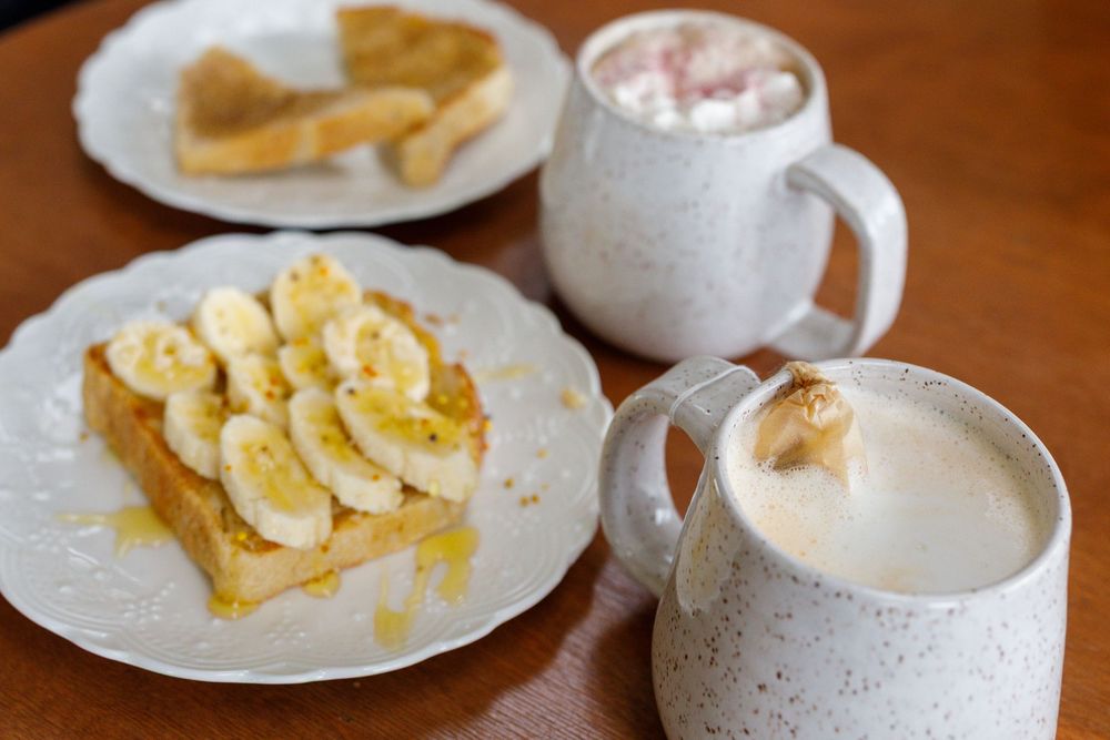 French toast topped with banana slices and honey on a white plate, served with two mugs of hot cocoa topped with marshmallows.