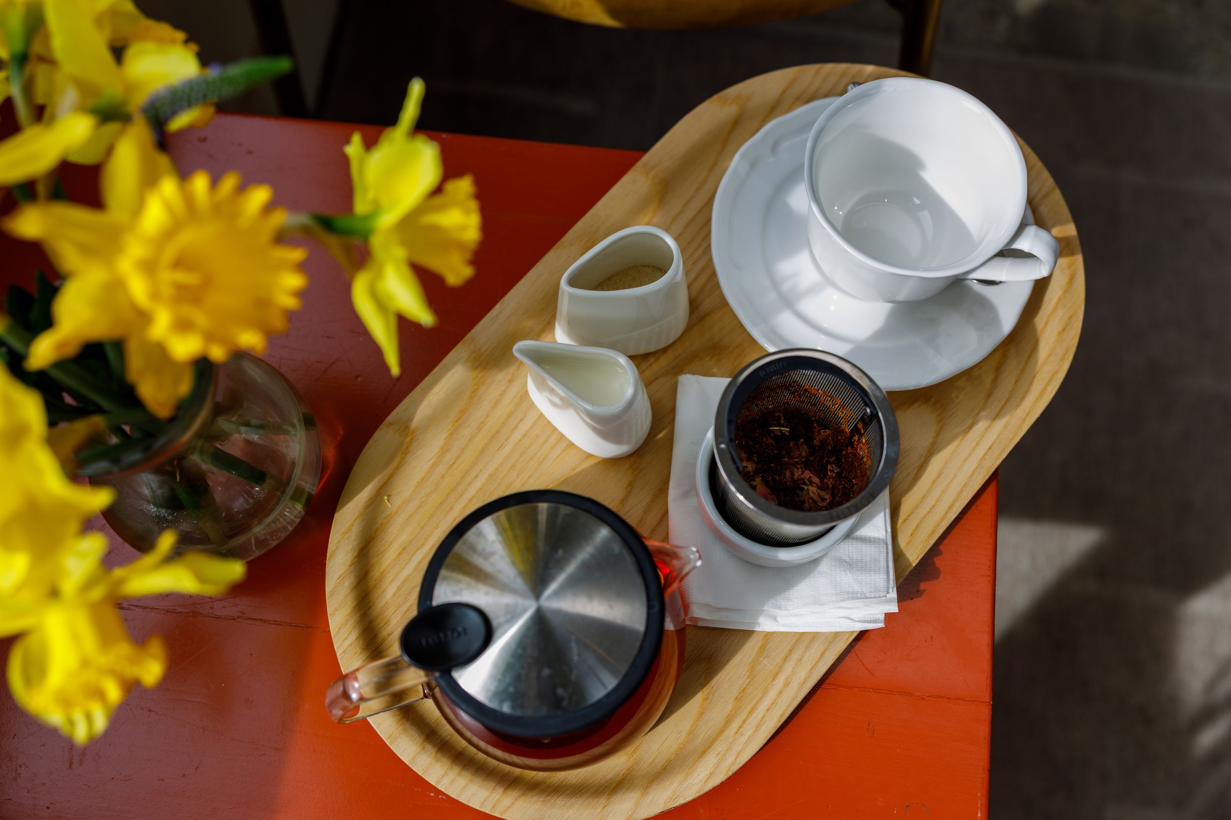 Overhead view of a wooden tray with white cup, creamer, and tea infuser on a red table beside yellow daffodils.
