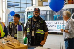 Workers in safety vests smile at an indoor transit facility event with blue balloons and a bus in the background.