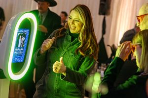 Woman in green jacket gives thumbs up at a neon-lit photo booth during an indoor event.