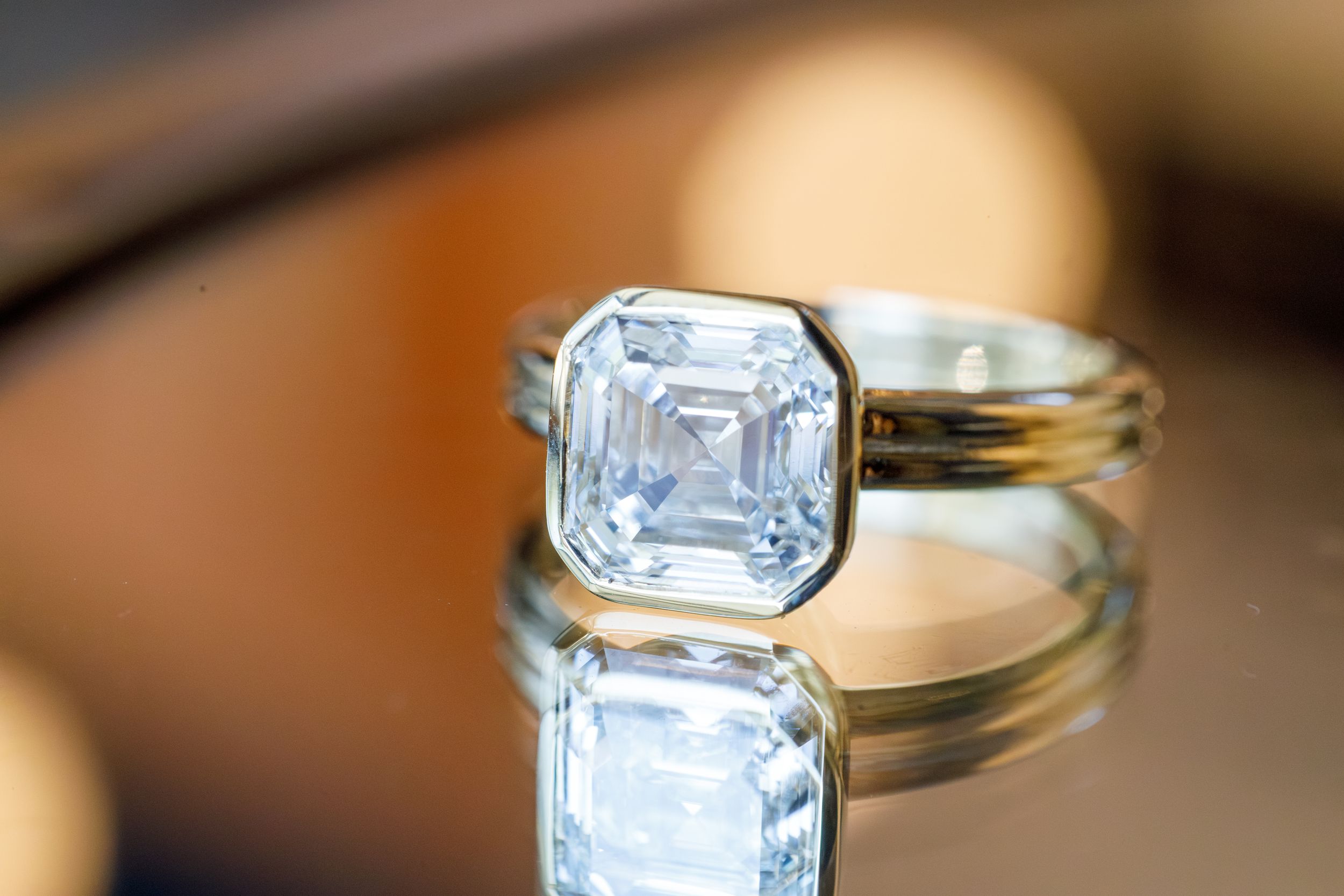 Elegant asscher-cut diamond solitaire ring in bezel setting on reflective gold surface.