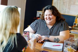 Two women smiling and talking at a table, one with tattoos, in a bright office setting.