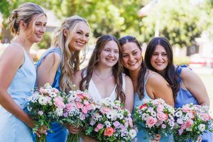 Bride in white dress with four bridesmaids in blue holding floral bouquets, smiling outdoors at a wedding.