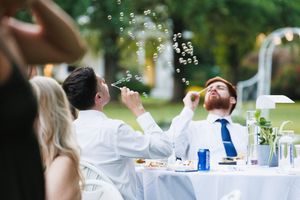 Two men blowing bubbles at an outdoor wedding reception table with white linens and greenery backdrop.