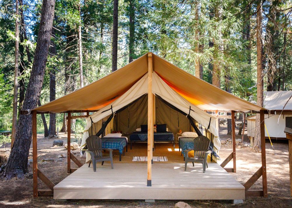 Luxury glamping tent with wooden deck, chairs, and table nestled among tall pine trees in a forest campsite.