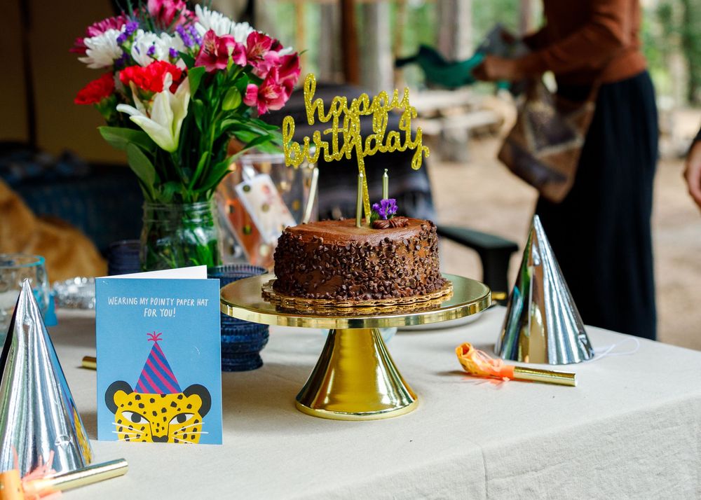 Birthday cake with happy birthday topper on gold stand, party hats, greeting card, and colorful flowers on white table.