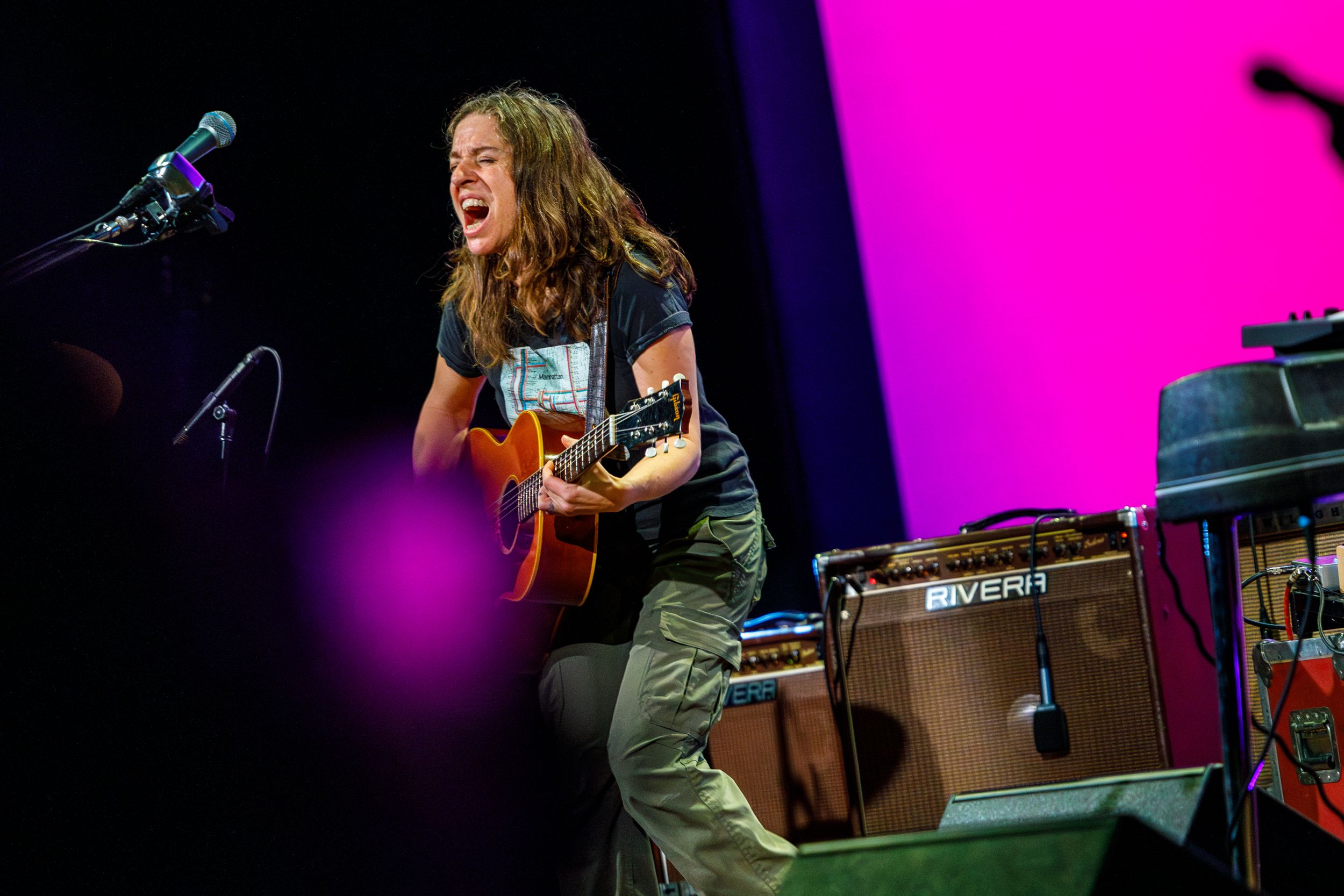 Young guitarist performing on stage with acoustic guitar, singing passionately under pink stage lights at live concert.
