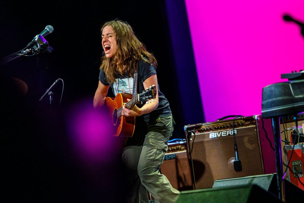 Young guitarist performing on stage with acoustic guitar, singing passionately under pink stage lights at live concert.