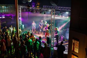 Live band performing on a lit stage with colorful lights while a crowd watches at an outdoor concert venue.