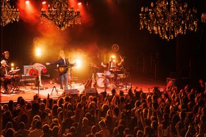 Live rock band performing on stage with chandeliers, dramatic red lighting, and an enthusiastic crowd.