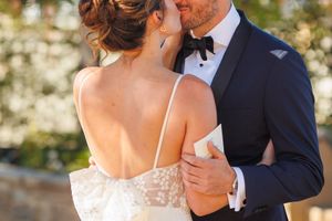 Bride and groom sharing a romantic kiss outdoors, wearing elegant wedding attire with lace gown and navy tuxedo.