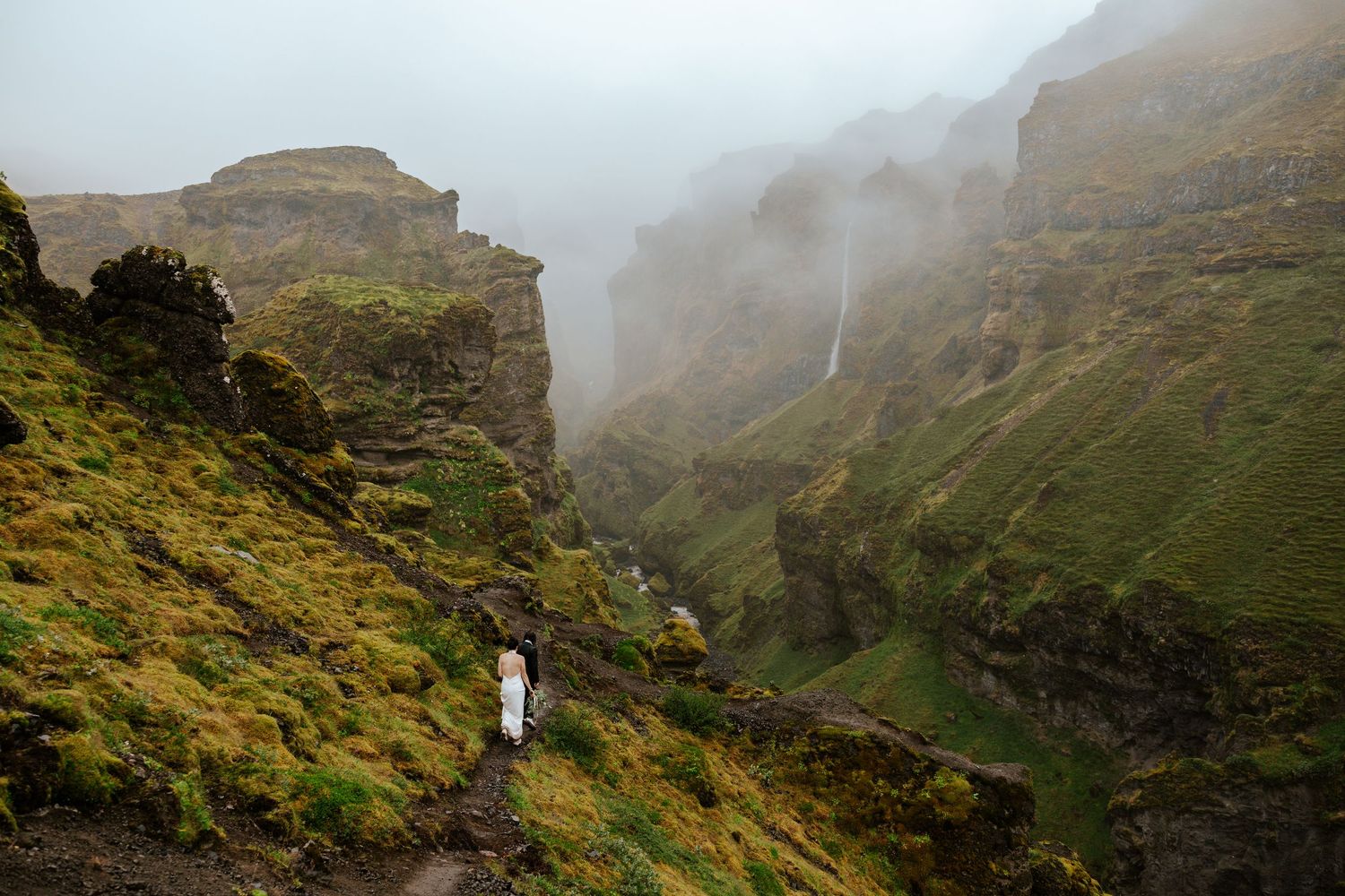 eloping couple walking down a mountain trail with a view of a waterfall and canyon at Mulagljufur Canyon during their Iceland elopement