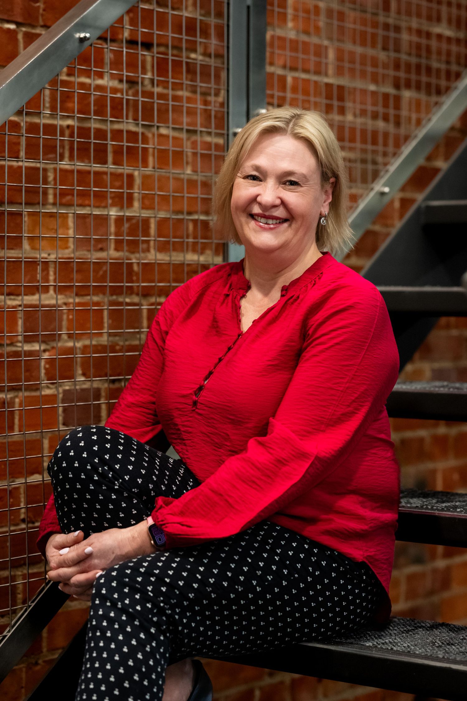 Smiling blonde woman in red blouse and patterned pants sitting on metal stairs against brick wall.
