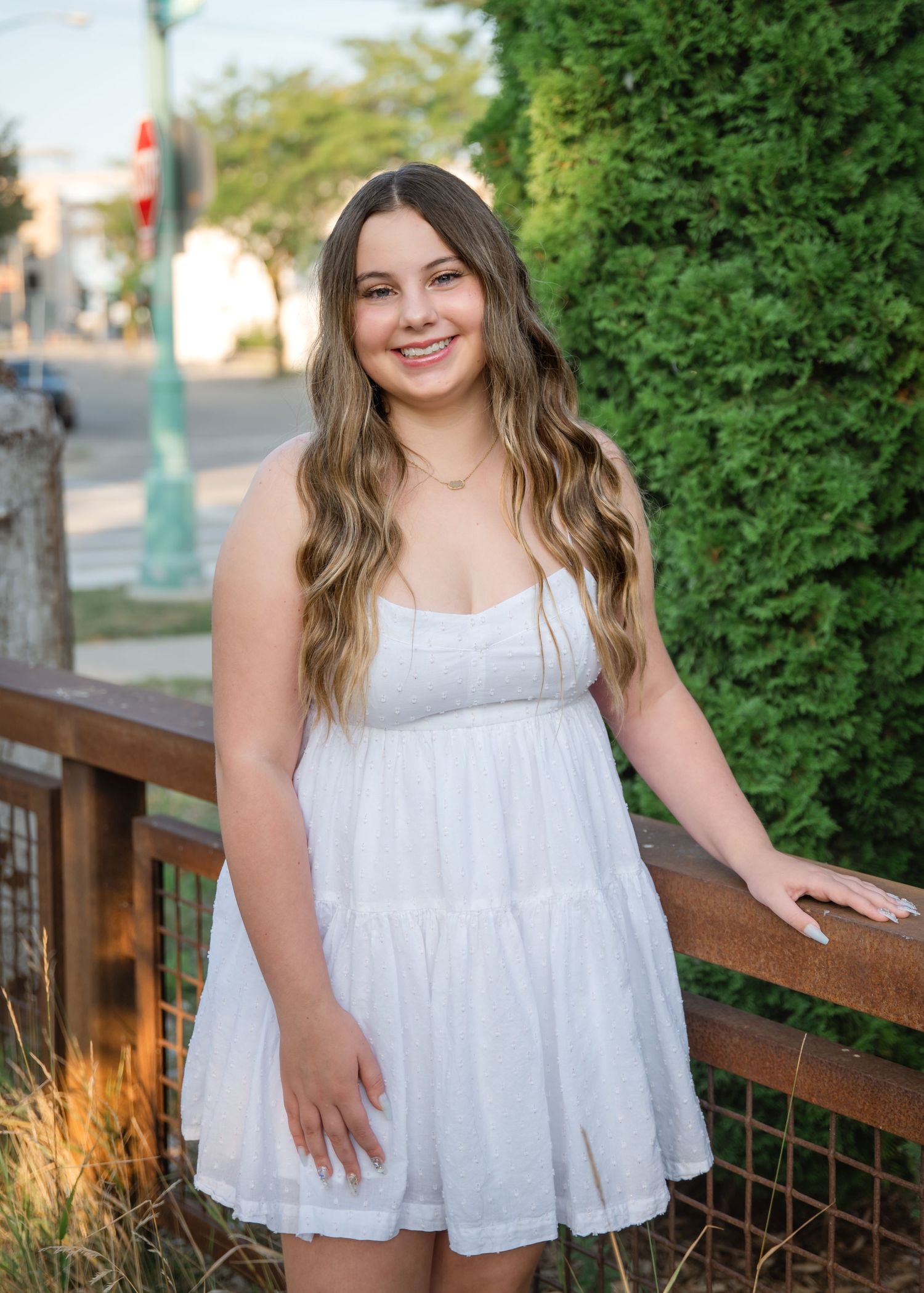 Young woman in white sundress smiling outdoors near wooden fence with lush green hedges in background.