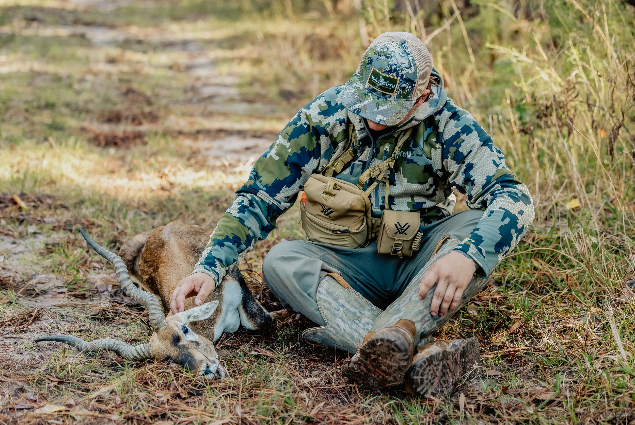 Hunter in camouflage gear examining harvested deer on ground in wooded area.