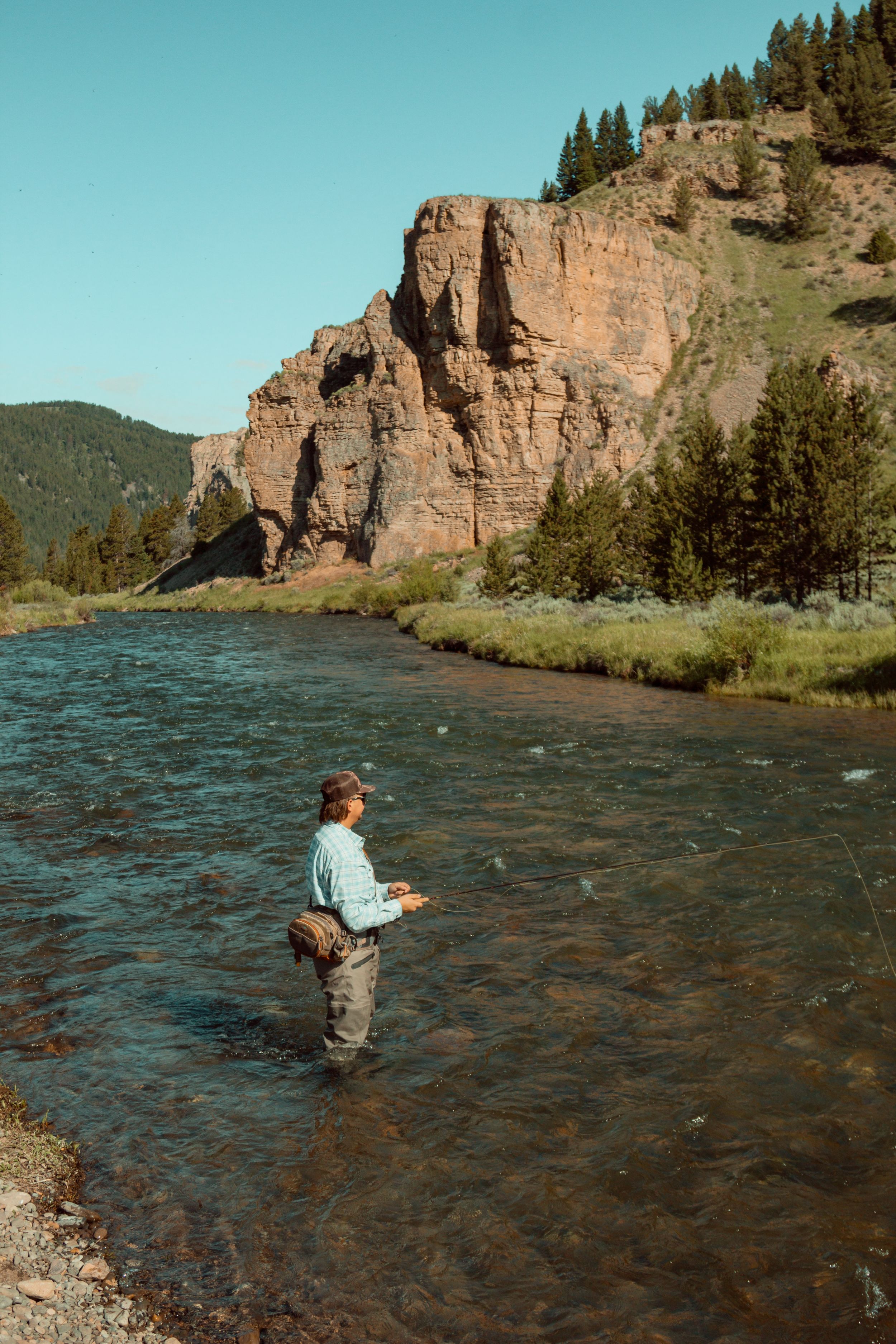 Fly fisherman wading in a river with dramatic red rock cliffs in background.