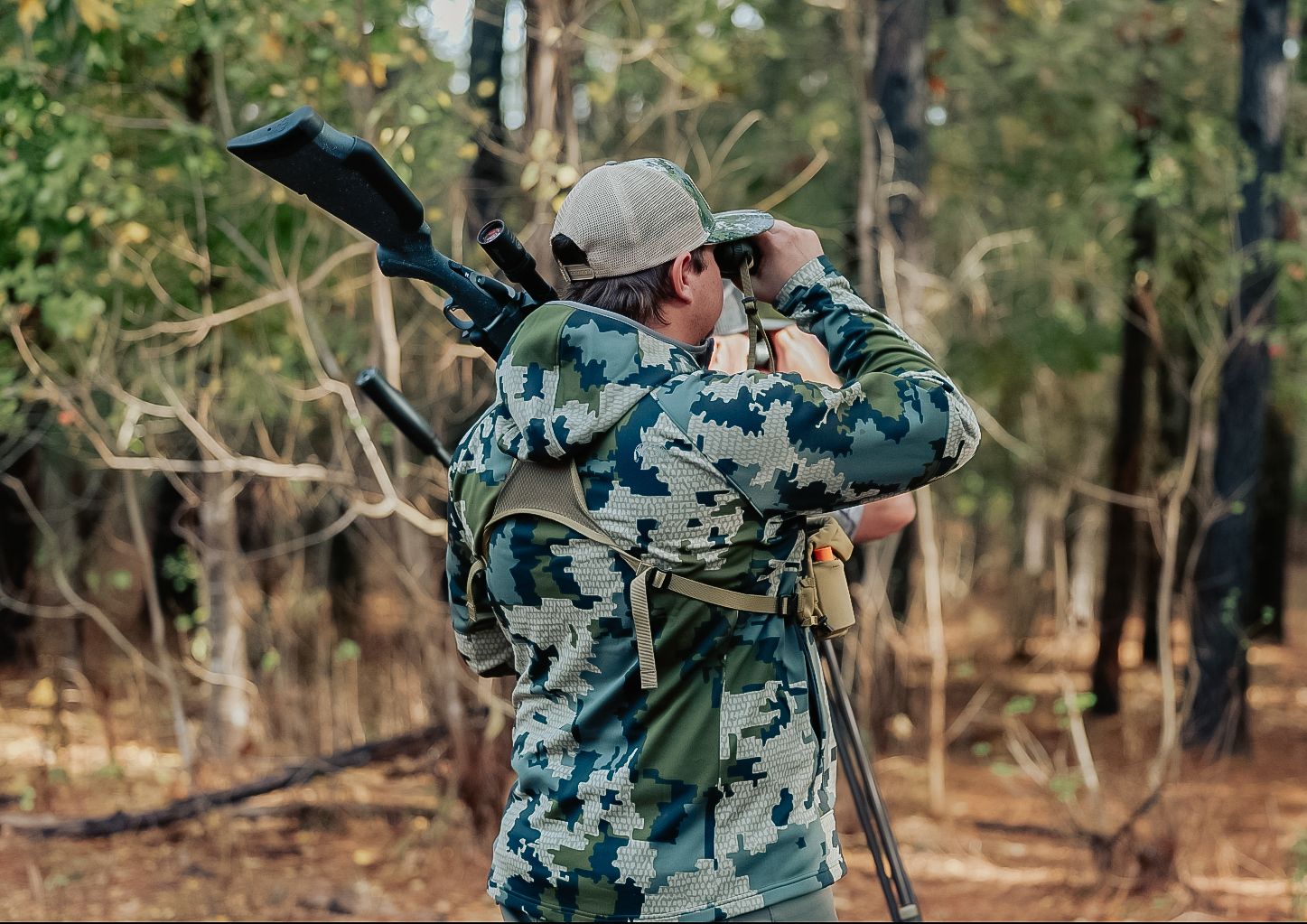 Hunter in camouflage jacket carrying rifle and using binoculars in forest.