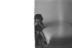 Black and white photo of a young child sitting on a bed, looking at the camera in a dimly lit room.