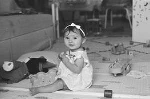 Black and white photo of a cute baby girl sitting on a play mat surrounded by toys, wearing a white dress and bow.
